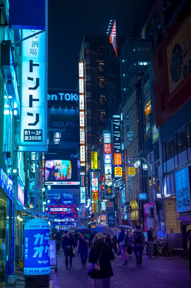 A narrow street at night in the rain, umbrellas moving between walls of neon